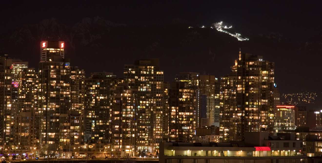 Downtown Vancouver at night, its towers lit, with the floodlit ski runs of Grouse Mountain on the dark ridge behind